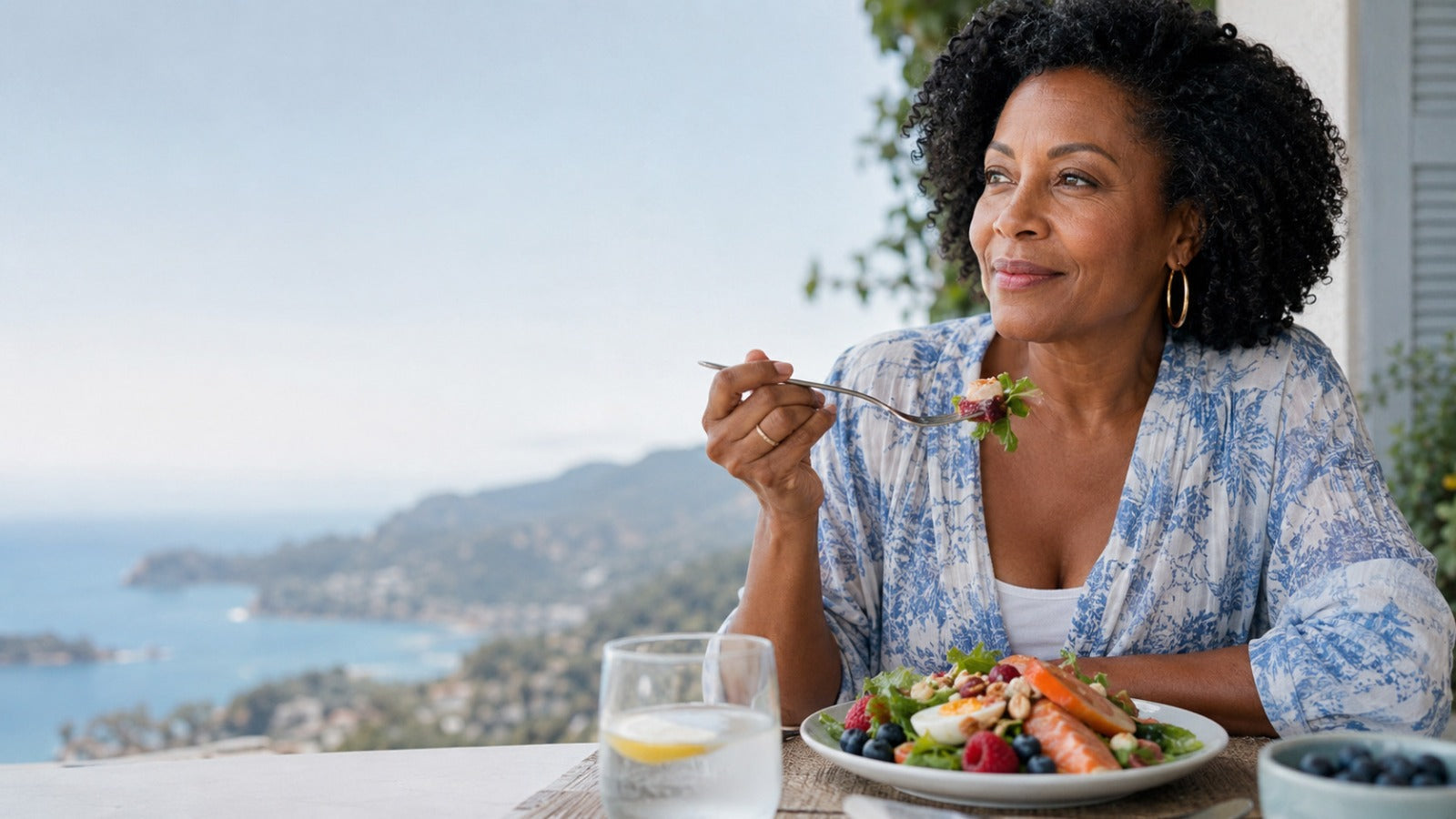 Black woman smiling and eating plate of colorful Mediterranean foods rich in lutein, zeaxanthin, antioxidants and omega-3 fats for women’s eye health, sitting in front of a window with beautiful coastal view