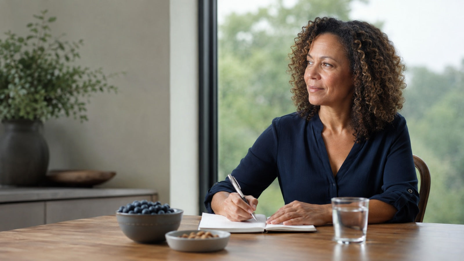 Midlife black woman journaling beside berries, walnuts and water in soft morning light, conveying renewed mental clarity.