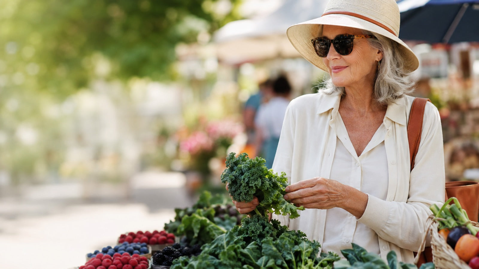 Active woman at a farmers market selecting leafy greens and berries while wearing UV-protective sunglasses and a hat.